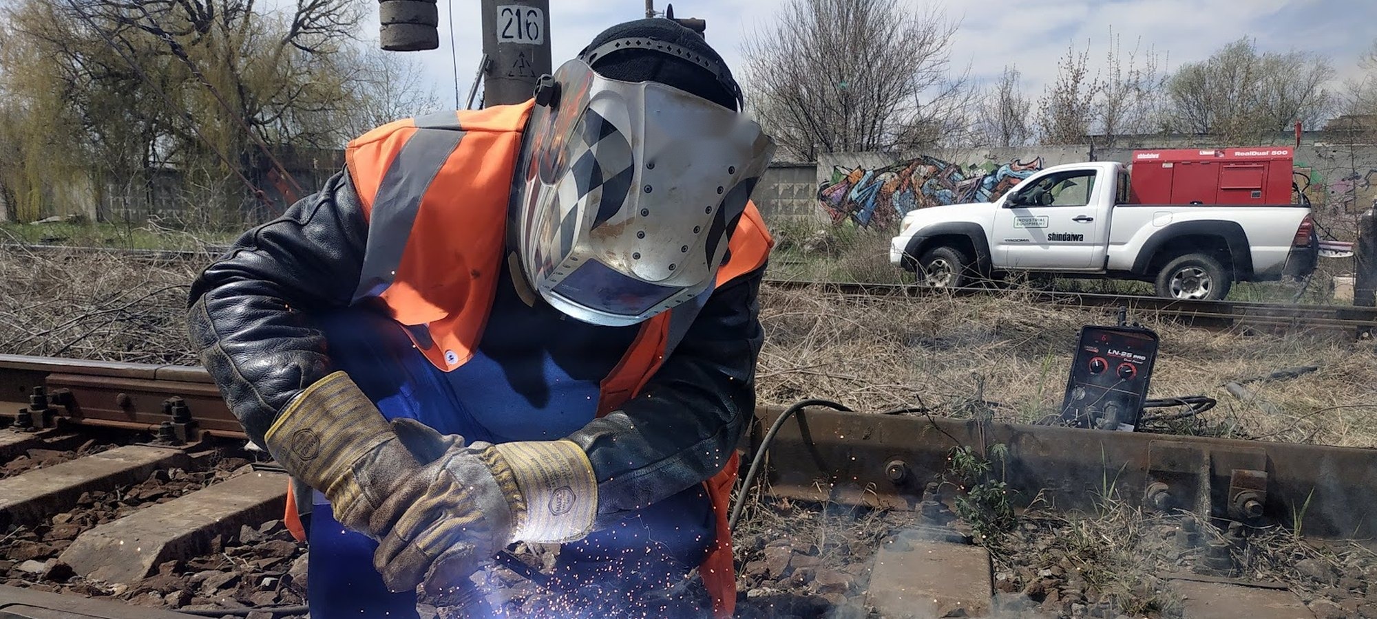 Svářeč opravuje kolejnici v terénu, v pozadí servisní pick-up se svařovacím agregátem SHINDAIWA